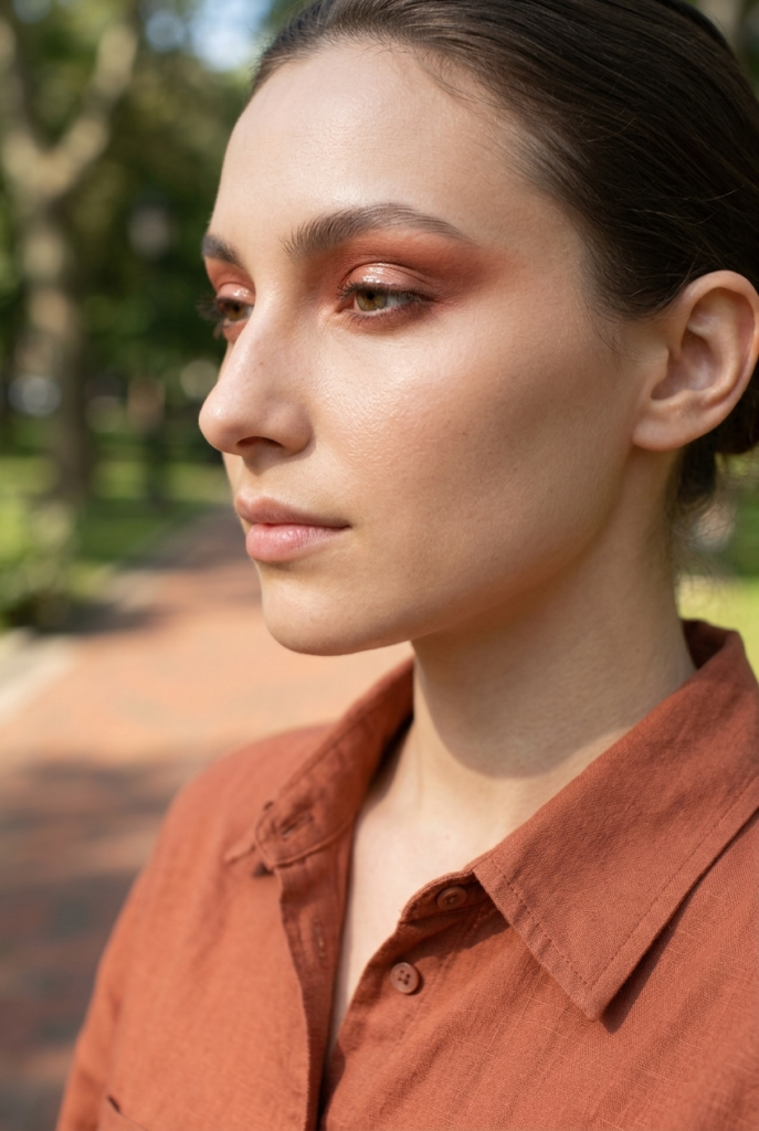Woman with a softened warm toned cut crease eye look in side profile in a park wearing a linen button-up shirt